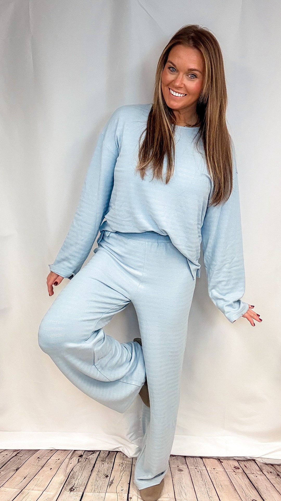 A woman modeling a light blue two-piece lounge set consisting of a long-sleeve top and matching wide-leg pants, standing against a white fabric backdrop on a wooden floor.