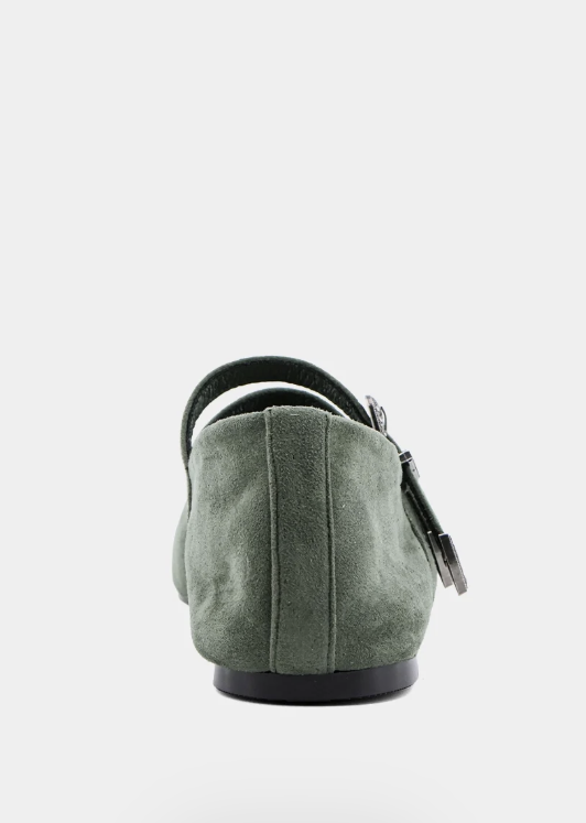 A single sage green suede flat shoe shown from the back, featuring a black sole and a small silver-tone metal charm on the side, set against a plain light gray background.