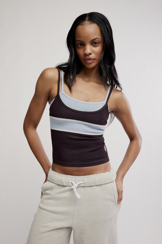 A woman modeling a color-blocked ribbed tank top with dark brown, light blue, and white horizontal stripes, paired with light beige sweatpants, against a plain light gray studio background.