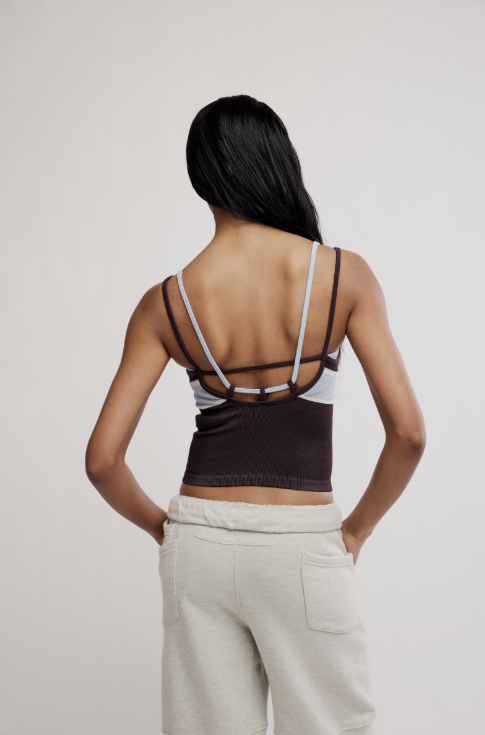 A woman shown from the back wearing a dark brown ribbed sports bra with thin black and light gray straps, paired with light beige sweatpants, against a plain light gray studio background.