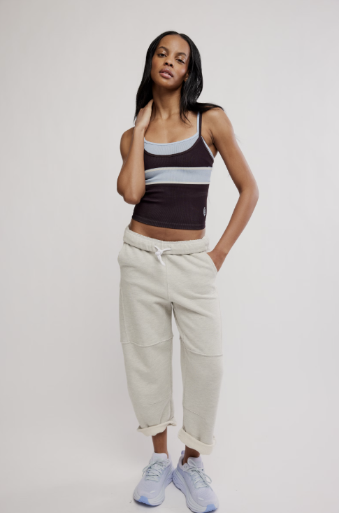 A model stands against a light gray studio background wearing a dark brown and light gray striped tank top, off-white cropped sweatpants, and light blue sneakers.