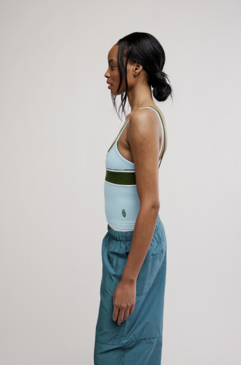 A woman in profile wearing a light blue ribbed tank top with a dark olive green horizontal band and matching light blue bottoms, photographed against a plain light gray studio background.