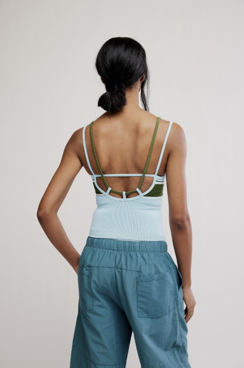 A woman shown from the back wearing a light blue ribbed tank top with olive green and white strappy details across the open back, paired with teal cargo pants, against a plain light gray studio background.