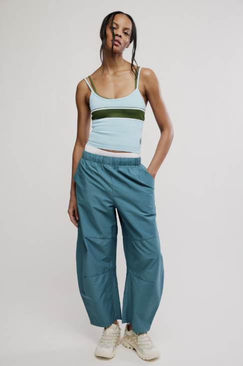 A model stands against a light gray studio background wearing loose teal-blue wide-leg pants, a light blue ribbed tank top with a dark green horizontal stripe, and off-white sneakers.