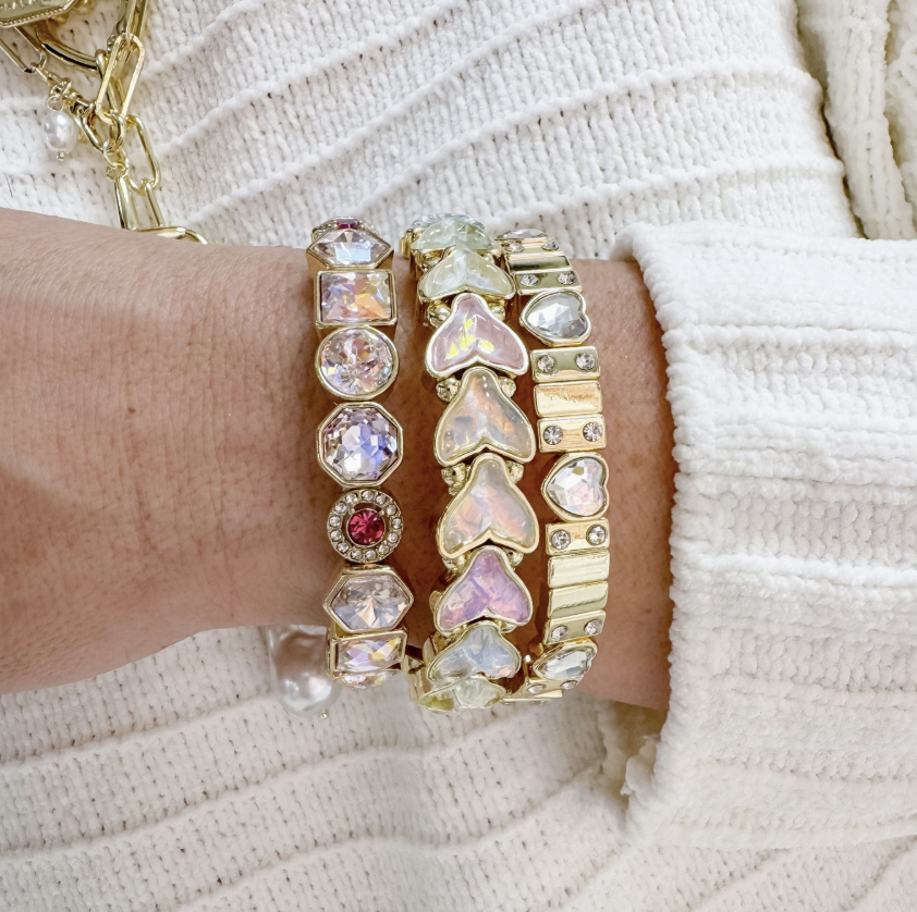 A close-up of a person's wrist wearing three stacked gold bracelets adorned with iridescent heart-shaped crystals, round clear stones, and one small pink gemstone, paired with a white knit sweater.