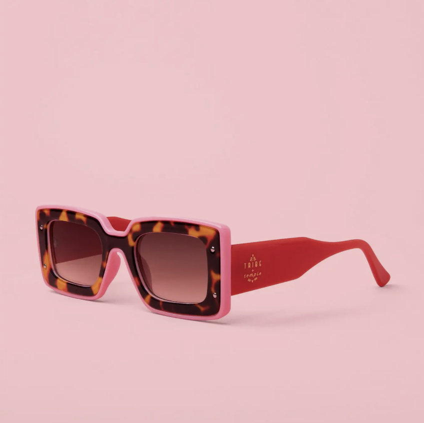 A pair of square sunglasses with a pink and tortoiseshell frame and red temples, displayed on a solid pink background.