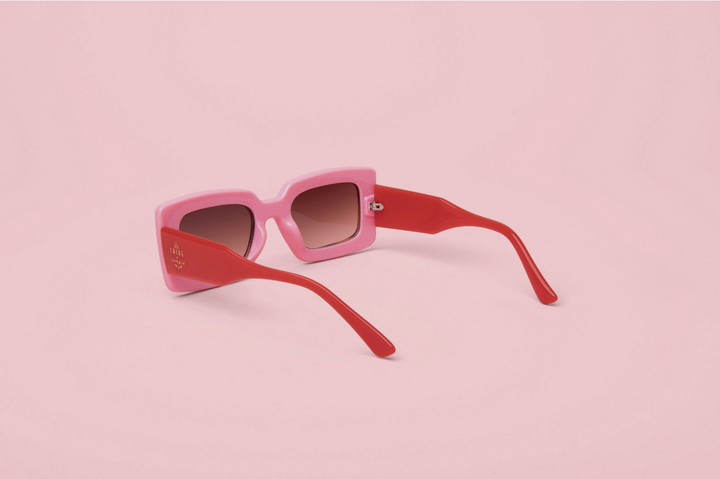 Pink and red cat-eye sunglasses with gradient brown lenses, displayed on a solid light pink background.