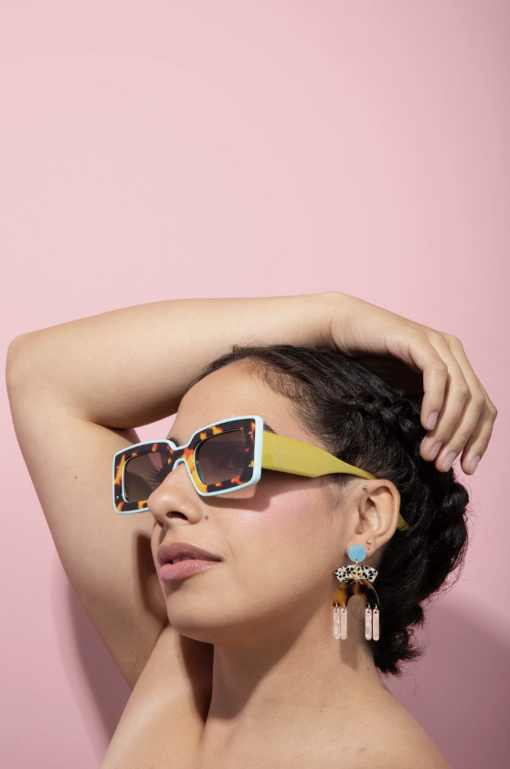 A woman wearing colorful square sunglasses with tortoiseshell-patterned lenses, yellow-green temples, and a light blue rim, posed against a solid pink background.