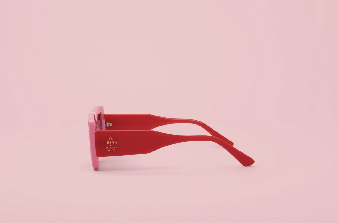A pair of square-frame sunglasses with translucent pink frames and red temples, shown in profile against a solid pink background.