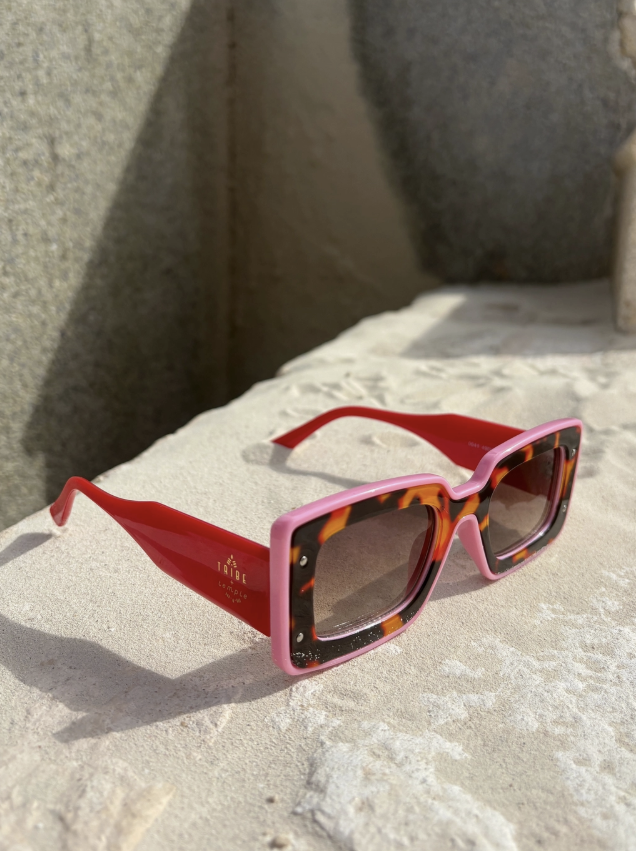 A pair of square sunglasses with a pink acetate frame, tortoiseshell inner rims, and bright red temples, resting on a textured light stone surface.