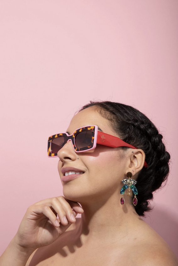 A woman wearing square sunglasses with a pink and tortoiseshell frame, posed against a solid pink background. She is also wearing dangling earrings.