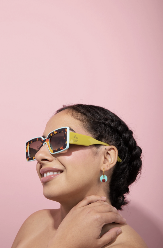 A woman with dark braided hair wearing colorful square sunglasses with a yellow temple and patterned frame, set against a solid pink background.