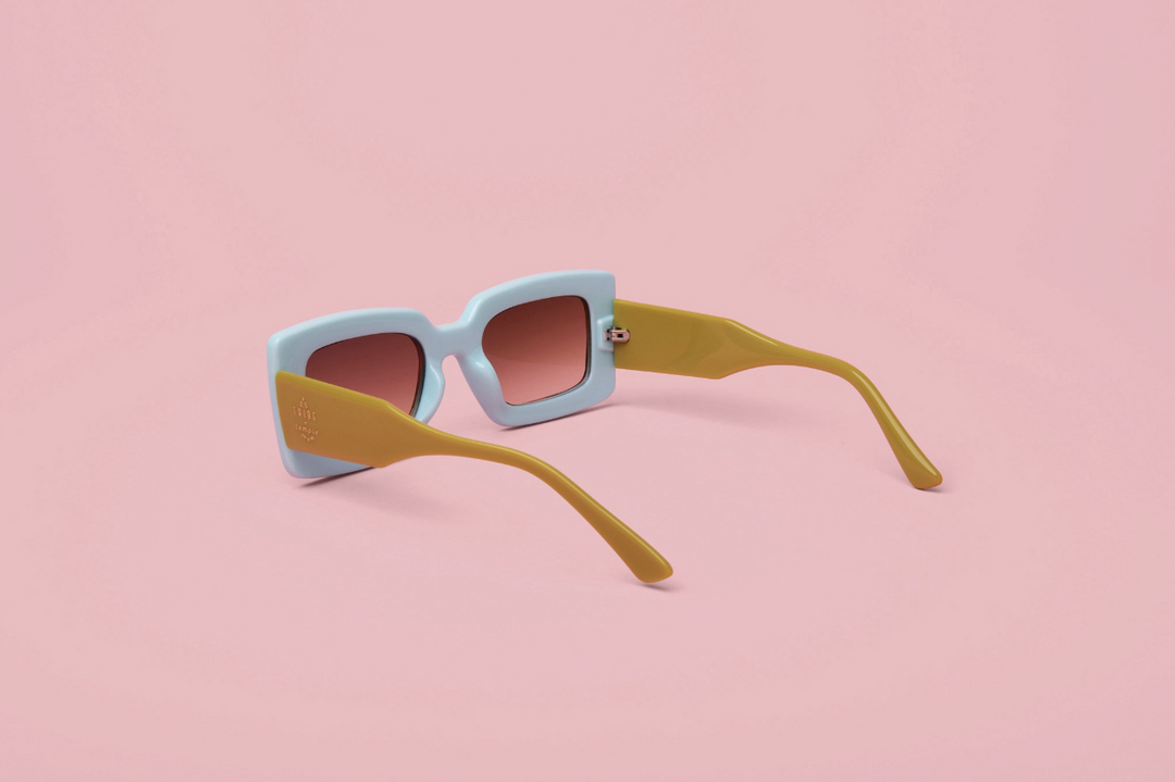 A pair of square sunglasses with a light blue front frame, mustard yellow arms, and pink-tinted lenses, displayed on a solid pink background.