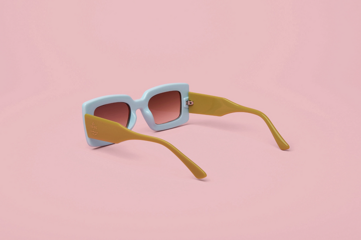 A pair of square sunglasses with a light blue front frame, mustard yellow arms, and pink-tinted lenses, displayed on a solid pink background.