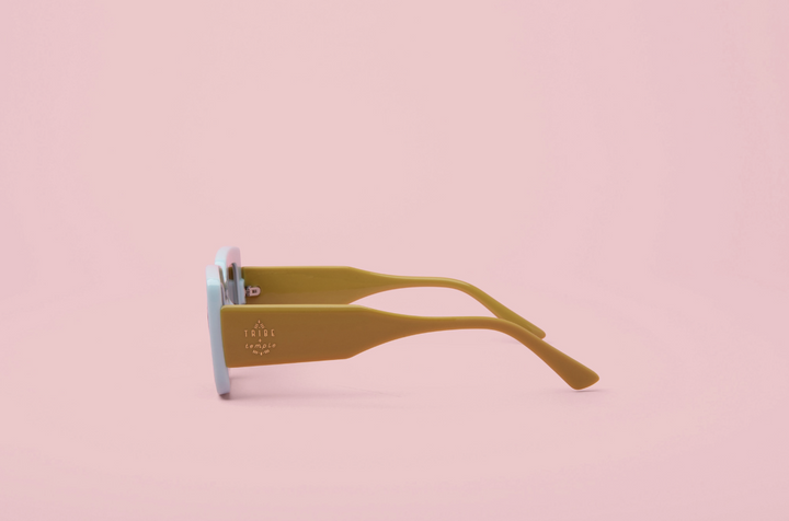 A pair of oversized square sunglasses with thick mustard yellow arms and light blue lenses, shown from a side profile on a solid pink background.