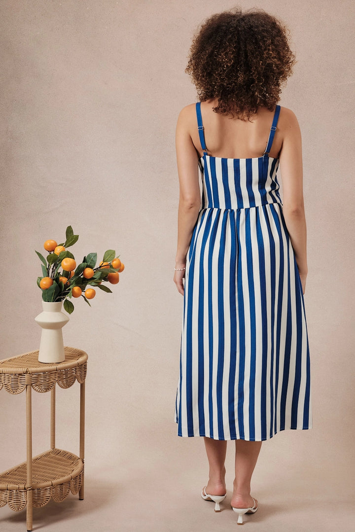 Woman wearing a blue and white striped dress standing in front of a beige wall with a small table and vase of oranges.