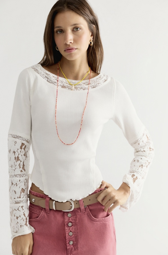 A woman modeling a white ribbed long-sleeve top with lace trim at the crew neckline and lace inserts on the sleeves, paired with pink jeans and layered necklaces against a plain light background.