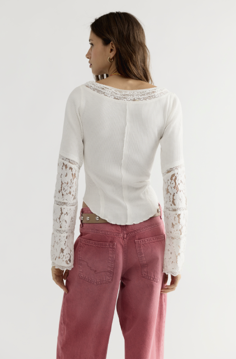A woman shown from the back wearing a white ribbed knit top with lace trim at the neckline and lace sleeves, paired with dusty rose jeans, against a plain light gray studio background.