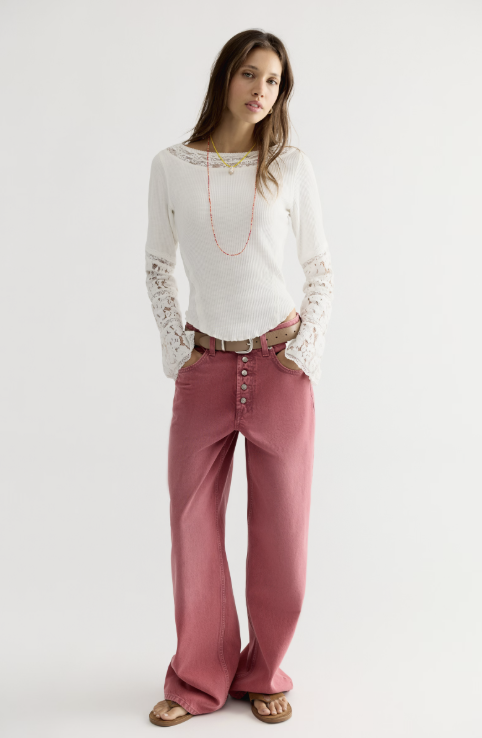 A woman modeling a white long-sleeve knit top with lace sleeves and matching white pants, styled with a brown belt, pinkish rose wide-leg jeans, and brown sandals against a light gray studio background.