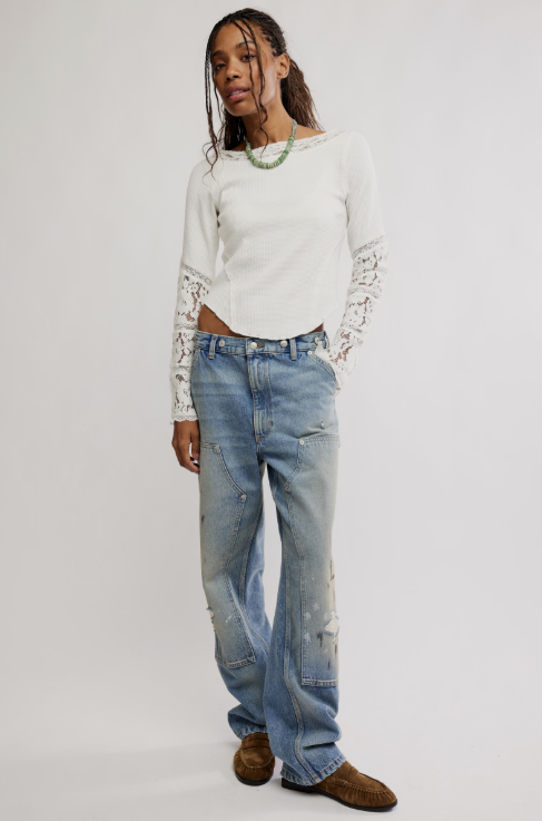 A model stands against a light gray studio background wearing a white long-sleeve top with lace sleeves, light-wash distressed blue jeans, and brown suede shoes.