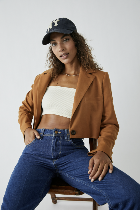 Woman wearing a brown blazer, white top, and blue jeans sitting on a chair against a white background