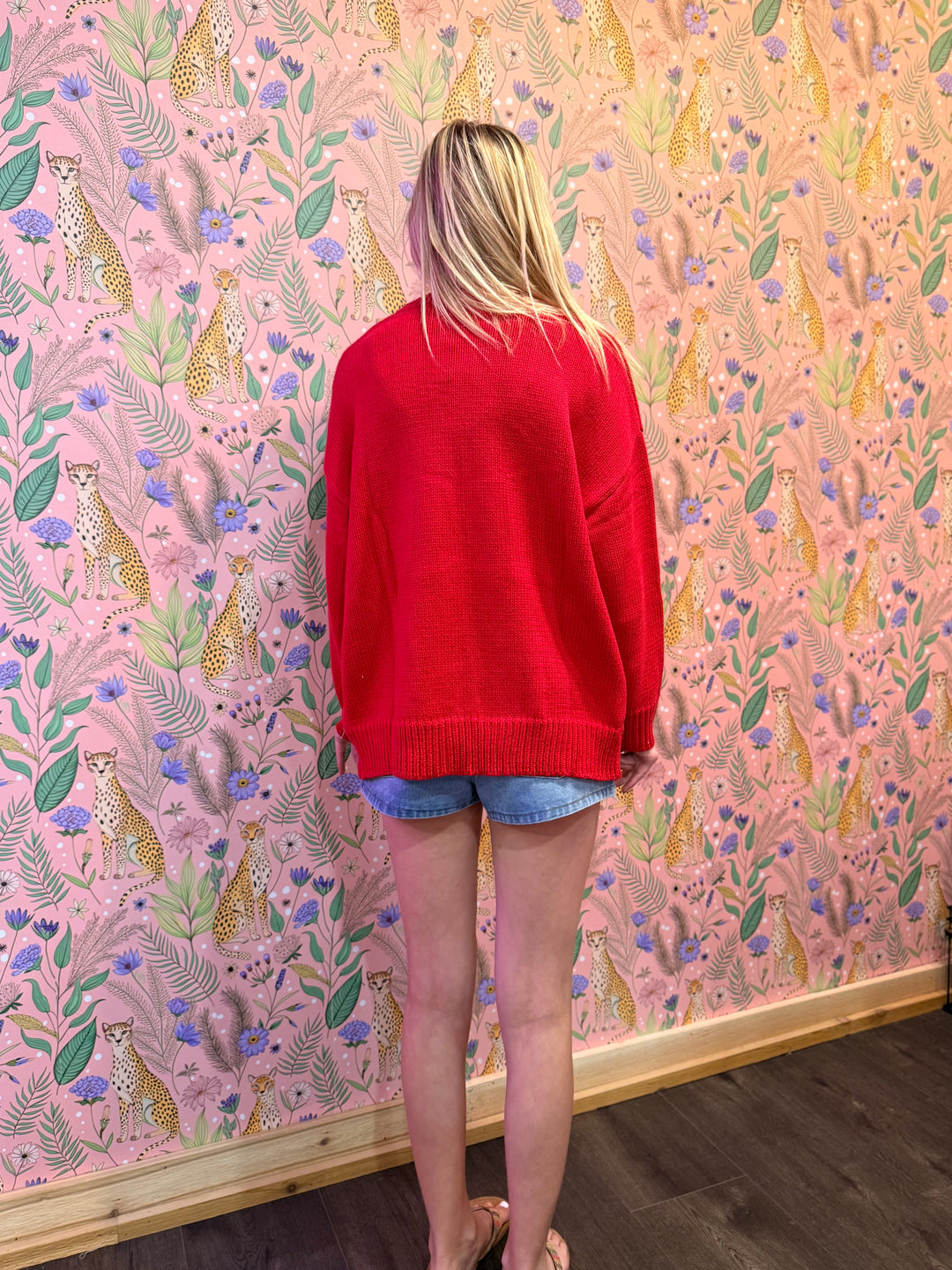 Person wearing a red sweater and denim shorts standing in front of a pink wall with animal and floral pattern.