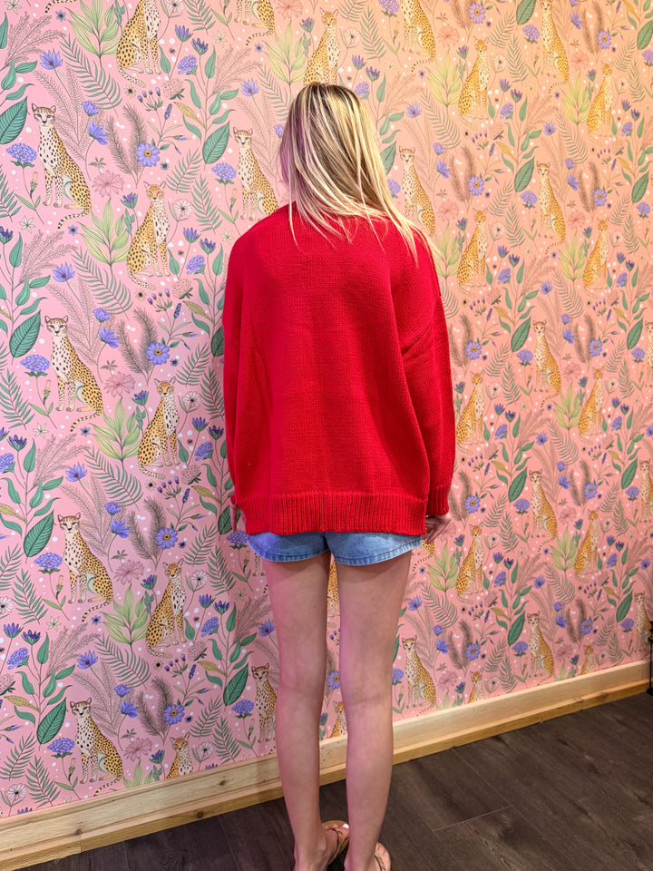 Person wearing a red sweater and denim shorts standing in front of a pink wall with animal and floral pattern.