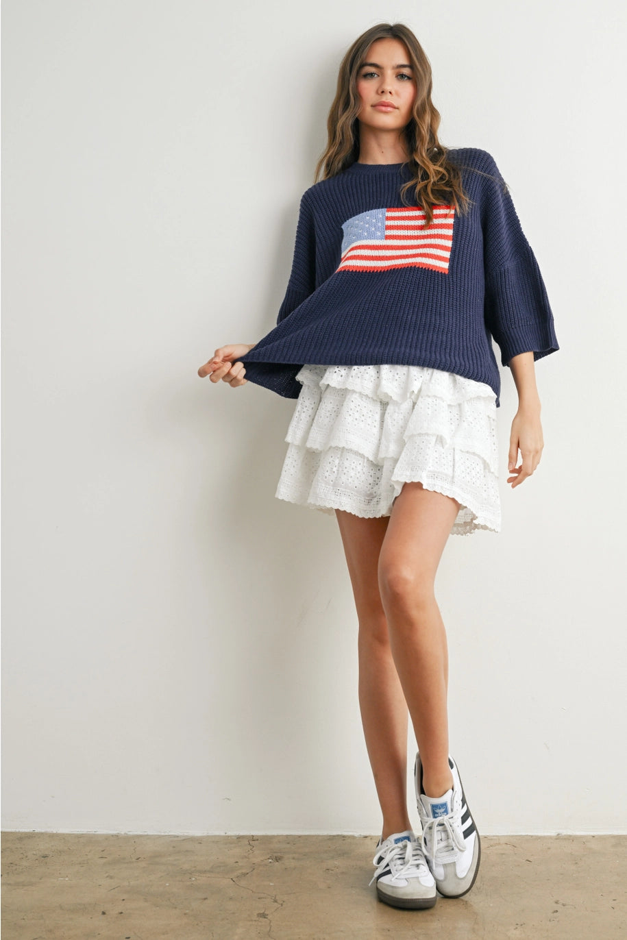 Woman wearing navy sweater with flag design and white skirt on plain background