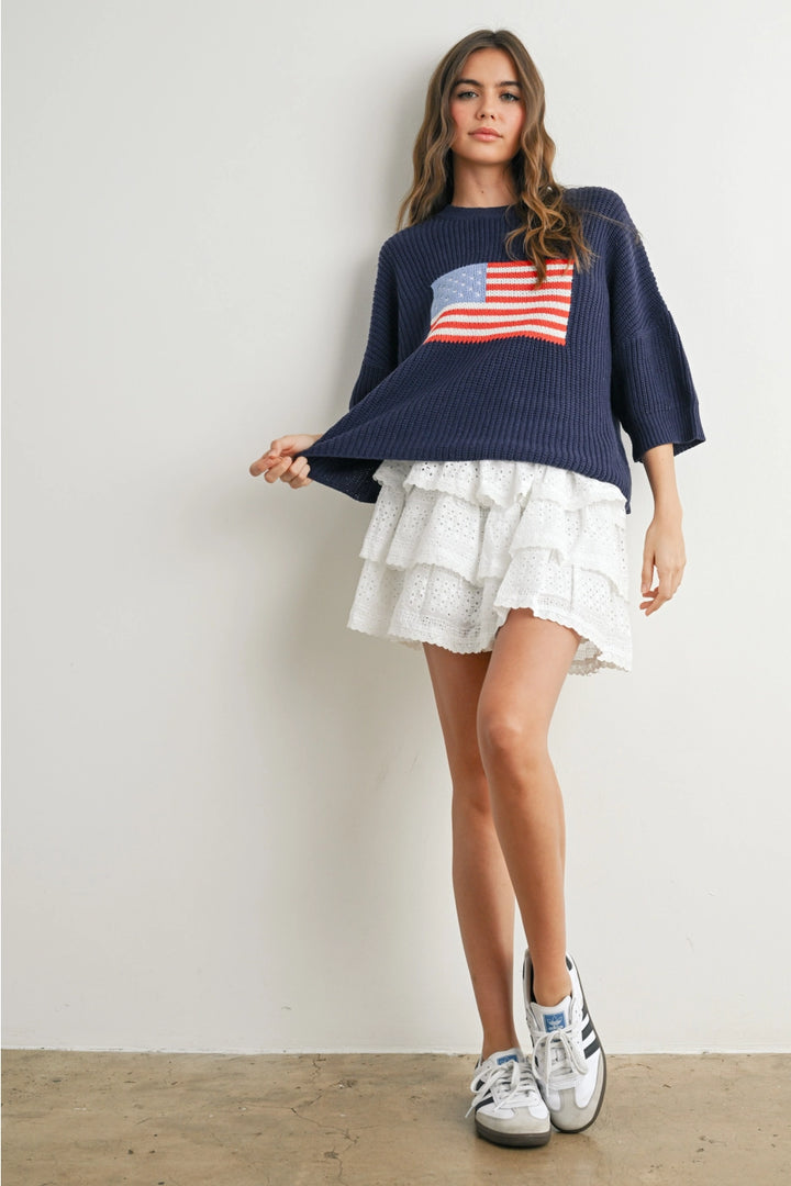 Woman wearing navy sweater with flag design and white skirt on plain background