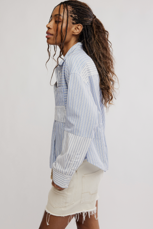 A woman shown in side profile wearing a light blue and white vertically striped button-up shirt with patch pockets, paired with a cream-colored denim mini skirt, against a plain light gray studio background.