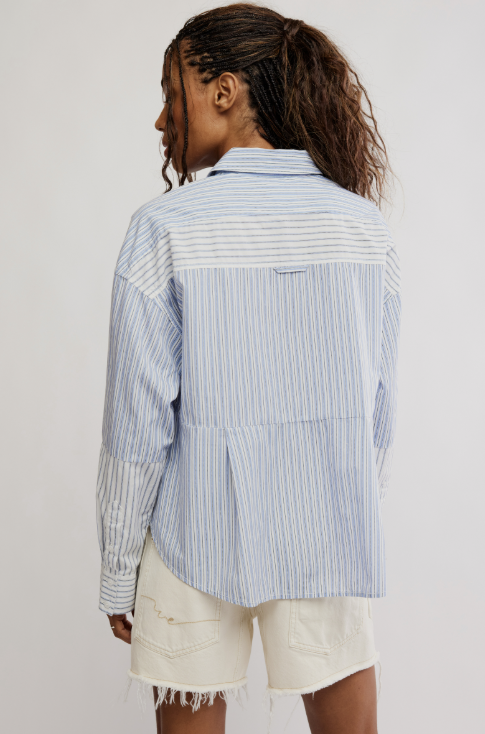 A woman shown from the back wearing a light blue and white striped patchwork button-up shirt and off-white denim shorts against a plain light gray studio background.