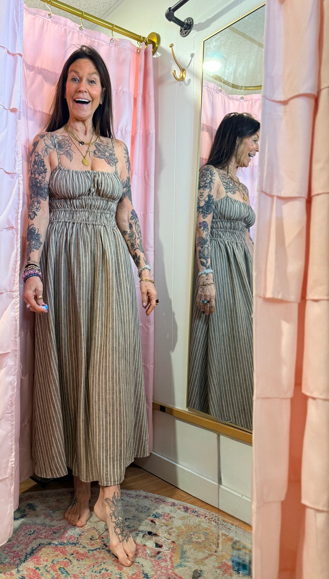Woman in a long dress standing in front of a mirror with a pink curtain in the background.