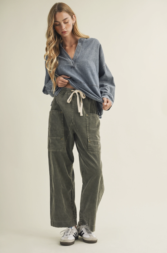 A woman modeling a pair of wide-leg olive green corduroy pants with a cream drawstring, styled with a blue-gray long-sleeve shirt and white sneakers against a plain beige studio background.