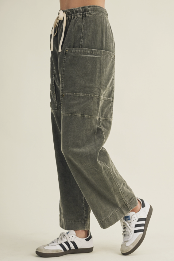 A person wearing loose, relaxed-fit corduroy cargo pants in a muted olive green color, paired with white low-top sneakers featuring black stripes, shown from the waist down against a plain light beige studio background.