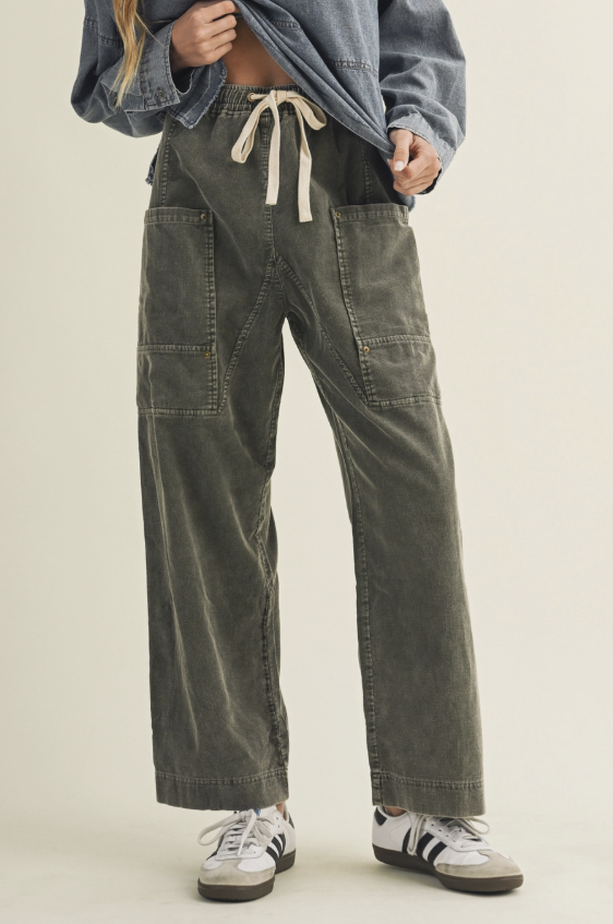 A person wearing wide-leg corduroy pants in an olive green color, styled with a blue denim shirt and white sneakers against a light beige background.