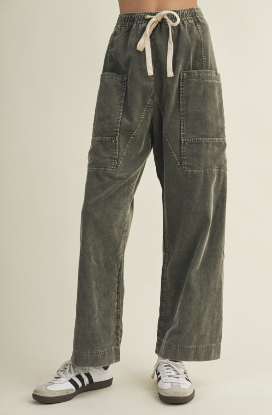 A person wearing wide-leg olive green corduroy pants with a cream drawstring waist, large front patch pockets, and cuffed hems, paired with white sneakers against a plain light beige studio background.