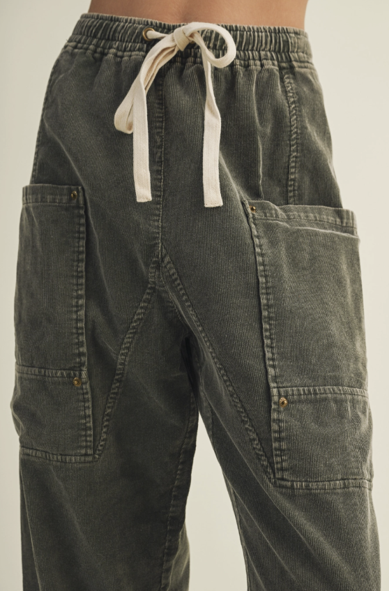 Close-up of a person wearing dark olive green cargo-style pants with a cream drawstring waist and large front patch pockets.