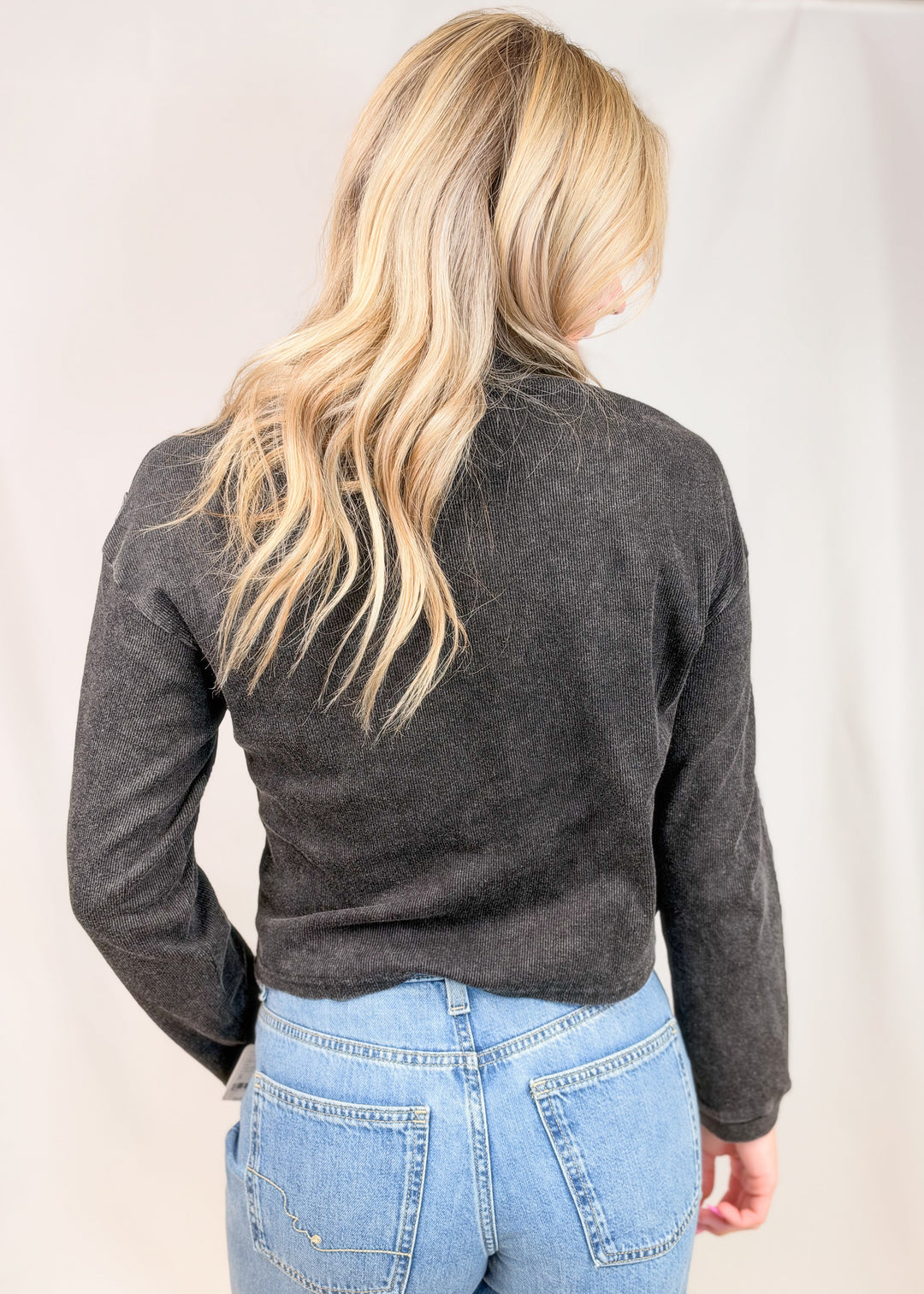 Woman shown from back view wearing black ribbed henley top with jeans on plain background