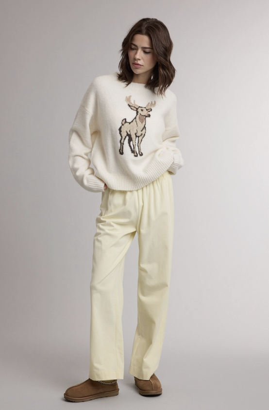 A woman stands against a light gray studio background wearing a cream knit sweater with a large deer graphic on the chest, matching cream wide-leg pants, and brown slippers.