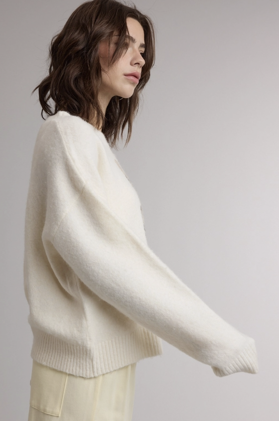 A woman in a side profile wearing a cream-colored, soft knit sweater and light cream pants against a plain light gray studio background.