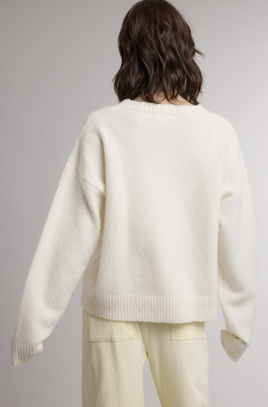 A model shown from the back wearing an off-white knit sweater and pale yellow pants against a neutral gray studio background.
