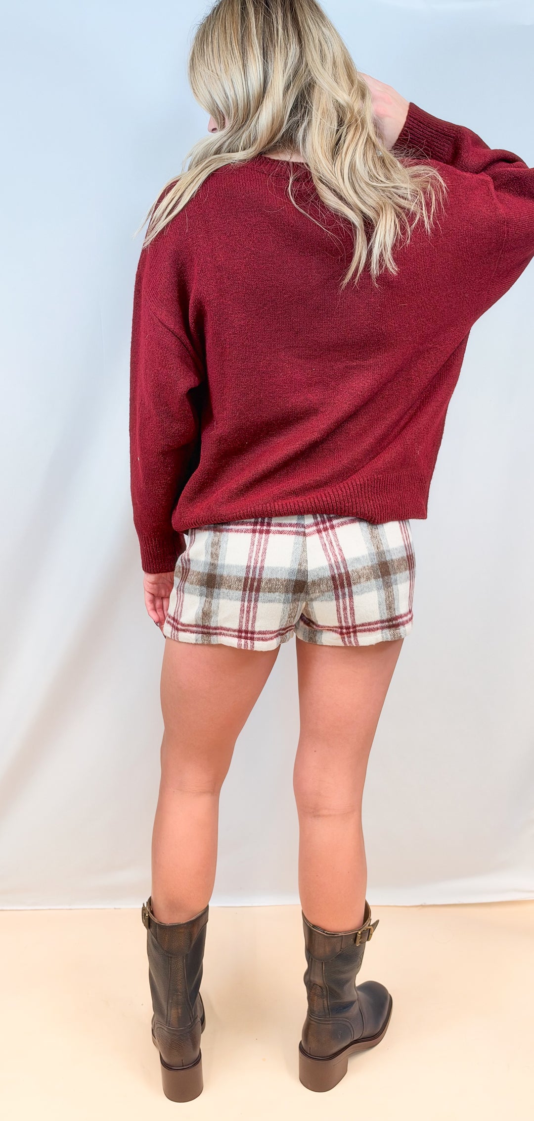 Woman shown from back view wearing burgundy sweater and plaid wool skort with boots on plain background