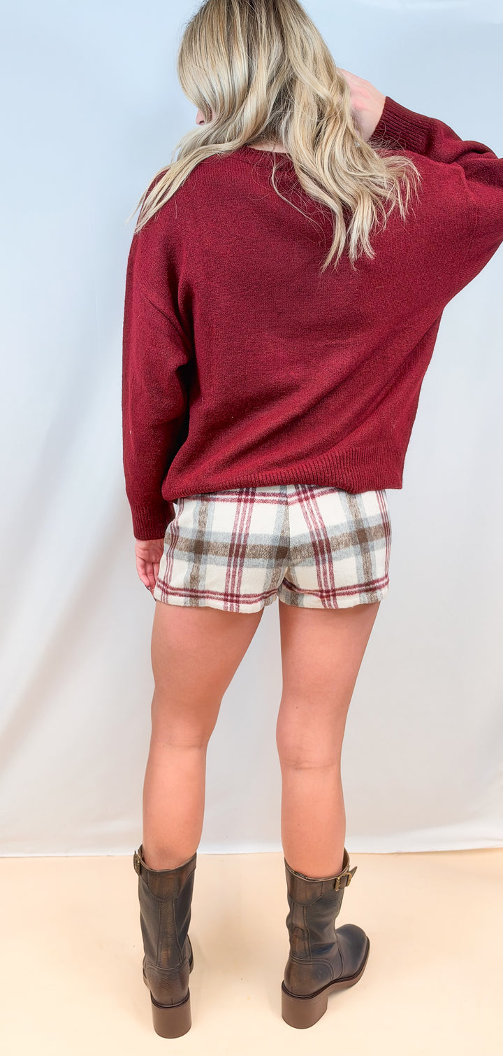 Woman shown from back view wearing burgundy sweater and plaid wool skort with boots on plain background