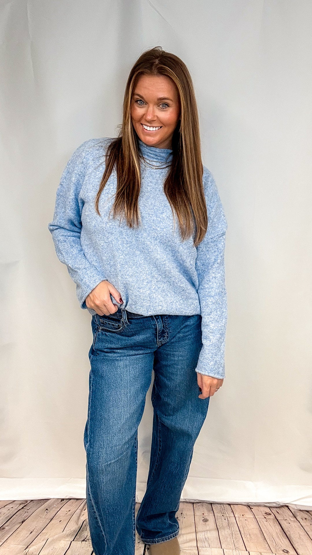 A woman modeling a light blue marled turtleneck sweater and dark blue jeans, standing against a white fabric backdrop on a wooden floor.