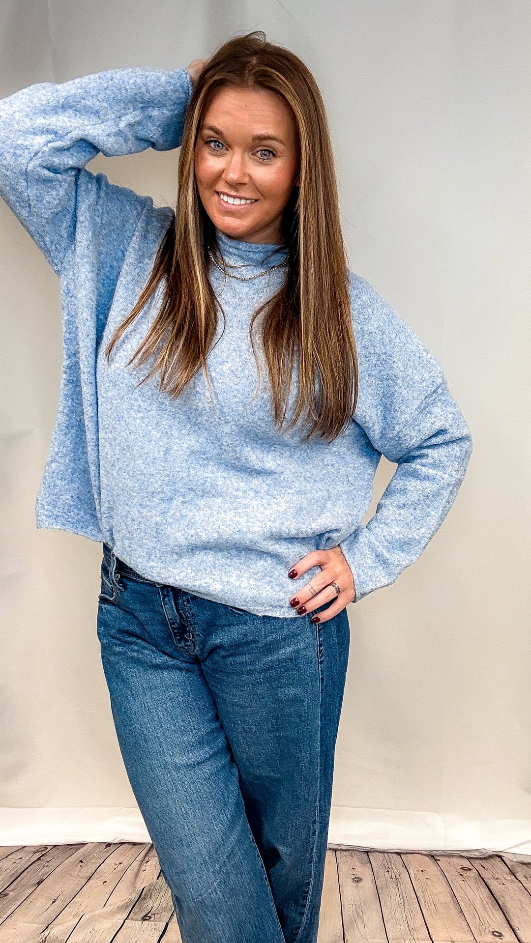 A woman modeling a light blue heathered knit turtleneck sweater and blue jeans, posing against a light gray studio backdrop with a wooden floor visible at the bottom.