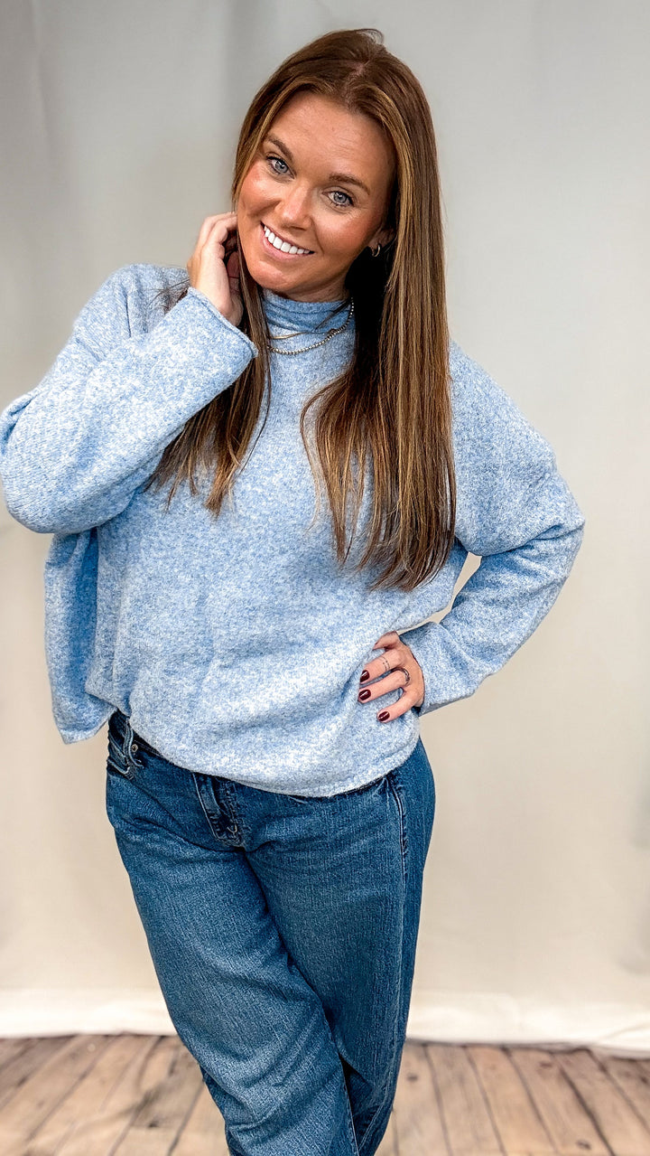 A woman modeling a light blue heathered turtleneck sweater and blue jeans against a neutral studio backdrop.