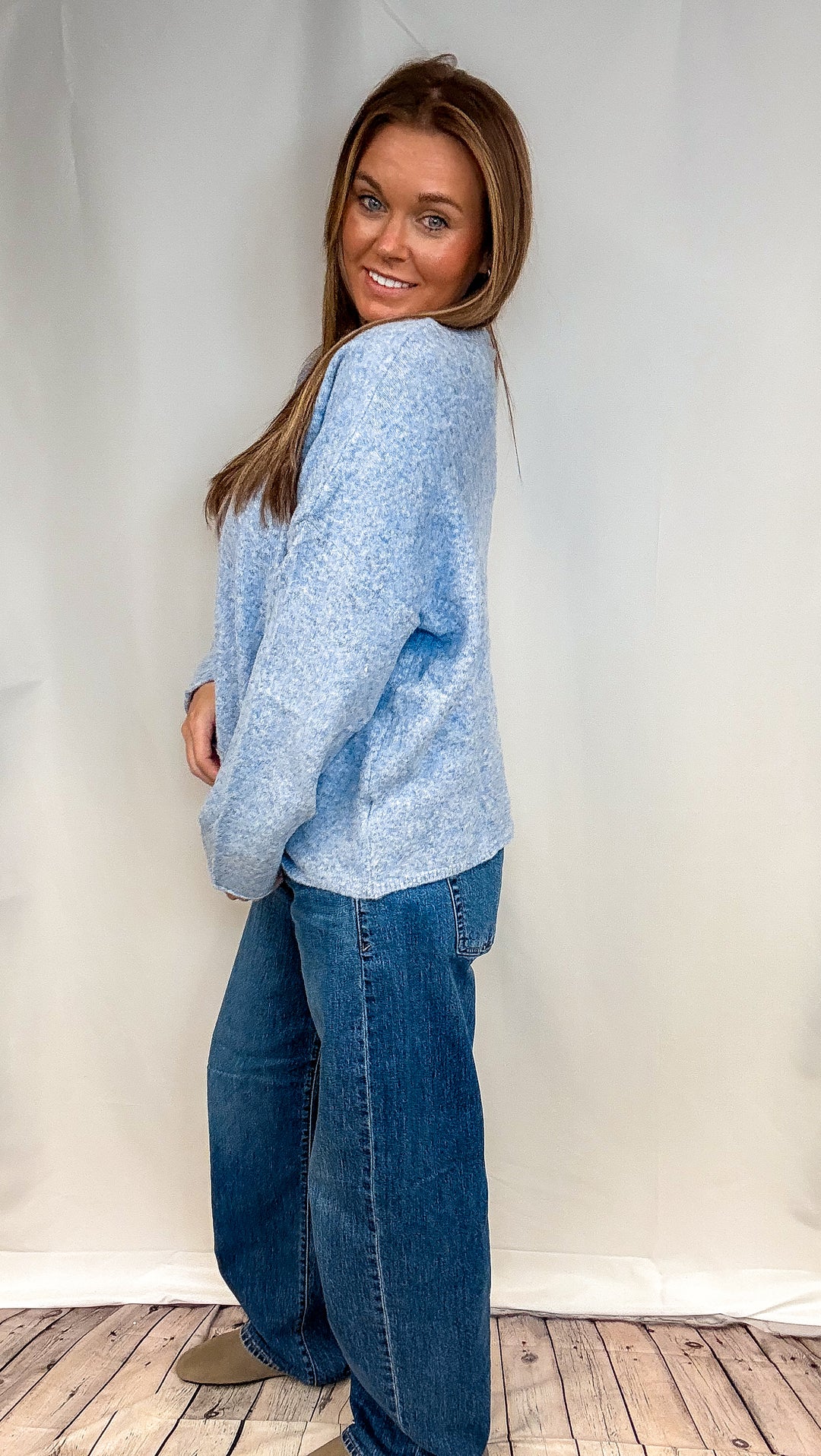 A woman wearing a light blue fuzzy knit sweater and blue jeans, standing in front of a white studio backdrop with a wood floor visible.