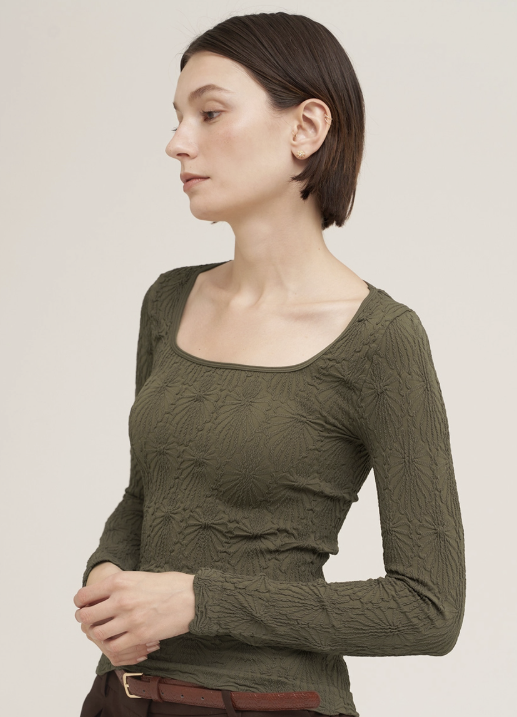 A woman wearing a textured olive green long-sleeve top with a square neckline, shown in a side profile against a plain light beige studio background.