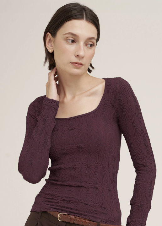 A woman wearing a textured, long-sleeve scoop-neck top in a deep plum purple color, paired with brown pants and a brown belt, against a plain light beige background.