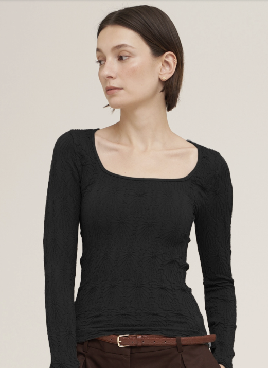 A woman wearing a fitted black long-sleeve top with a square neckline and subtle textured pattern, paired with brown pants and a brown belt, against a plain light beige studio background.